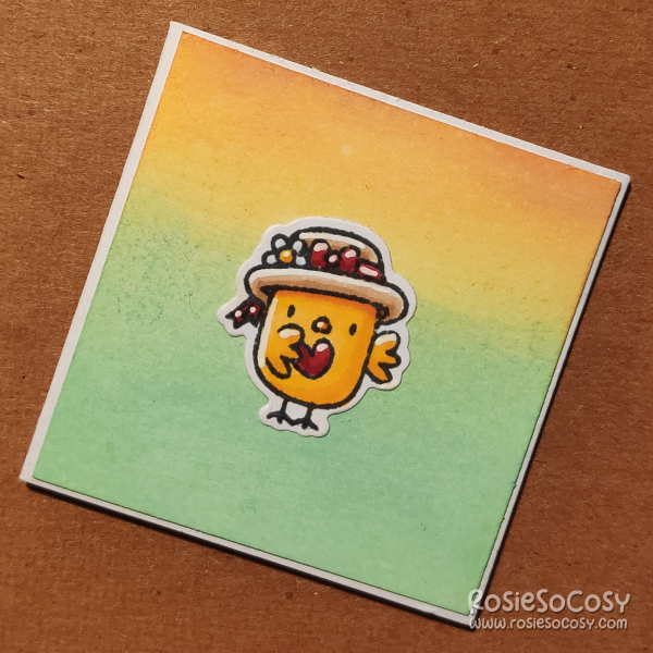 A inch sized card with a yellow to green gradient background. On the card is a tiny yellow chick with a cute floral hat, holding a tiny red heart.