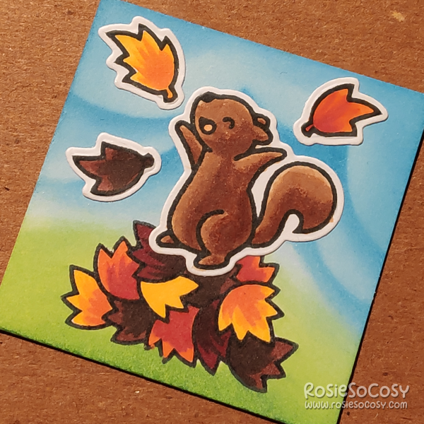 A tiny 2 inch card with a blue cloudy background, and a green grassy ground below, there is a overtly happy brown squirrel dancing atop a big pile of leaves, with a few of those leaves flying around.